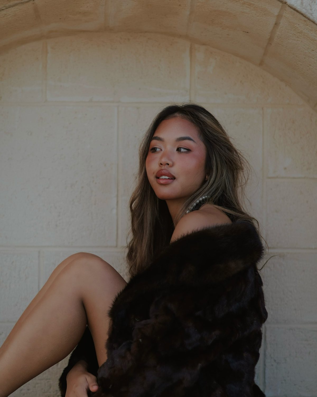 Kara Chaliaowai seated against a sandstone archway in a dark fur coat, looking off camera in daylight